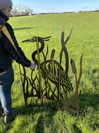 A person in a navy and yellow jacket is adjusting a rust-colored metal garden sculpture of a heron among reeds, set in a grassy field.