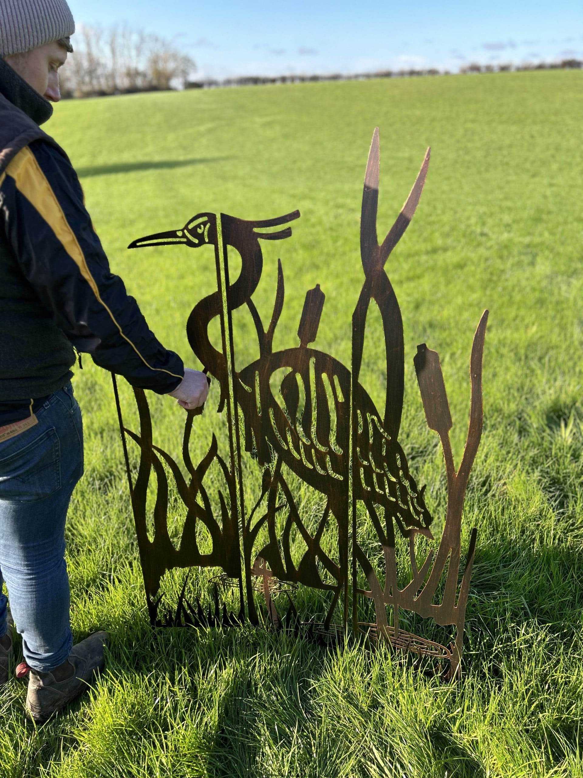 A person in a navy and yellow jacket is adjusting a rust-colored metal garden sculpture of a heron among reeds, set in a grassy field.