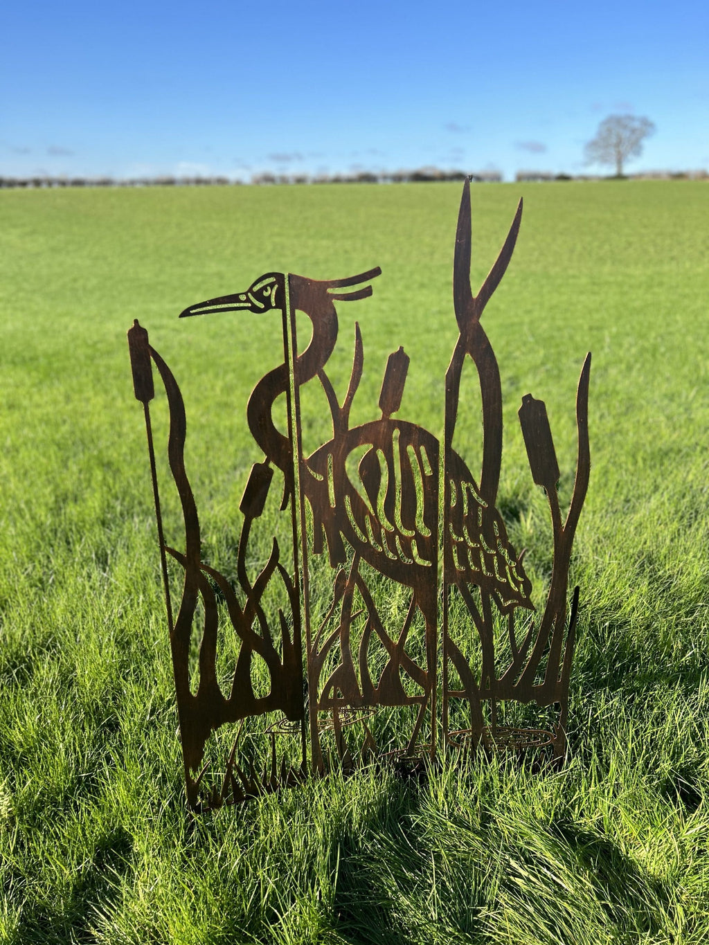 Rustic metal garden sculpture of a heron and reeds, displayed outdoors in a green grassy field under a clear blue sky.
