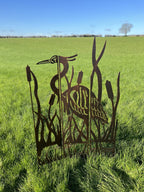 Rustic metal garden sculpture of a heron and reeds, displayed outdoors in a green grassy field under a clear blue sky.