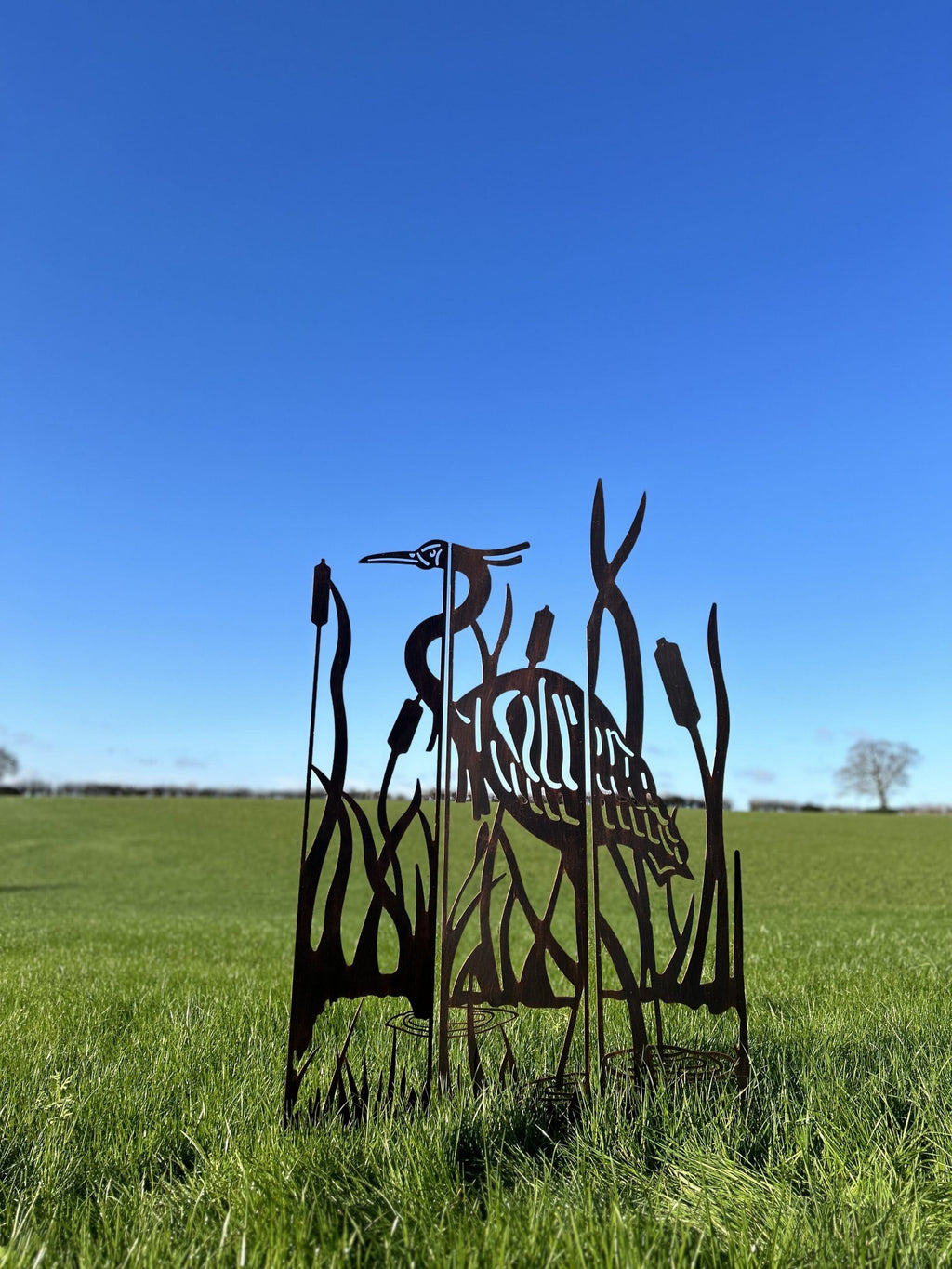A metal garden sculpture of a heron among reeds, shown outdoors in a grassy field under a clear blue sky.