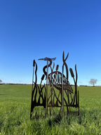 A metal garden sculpture of a heron among reeds, shown outdoors in a grassy field under a clear blue sky.