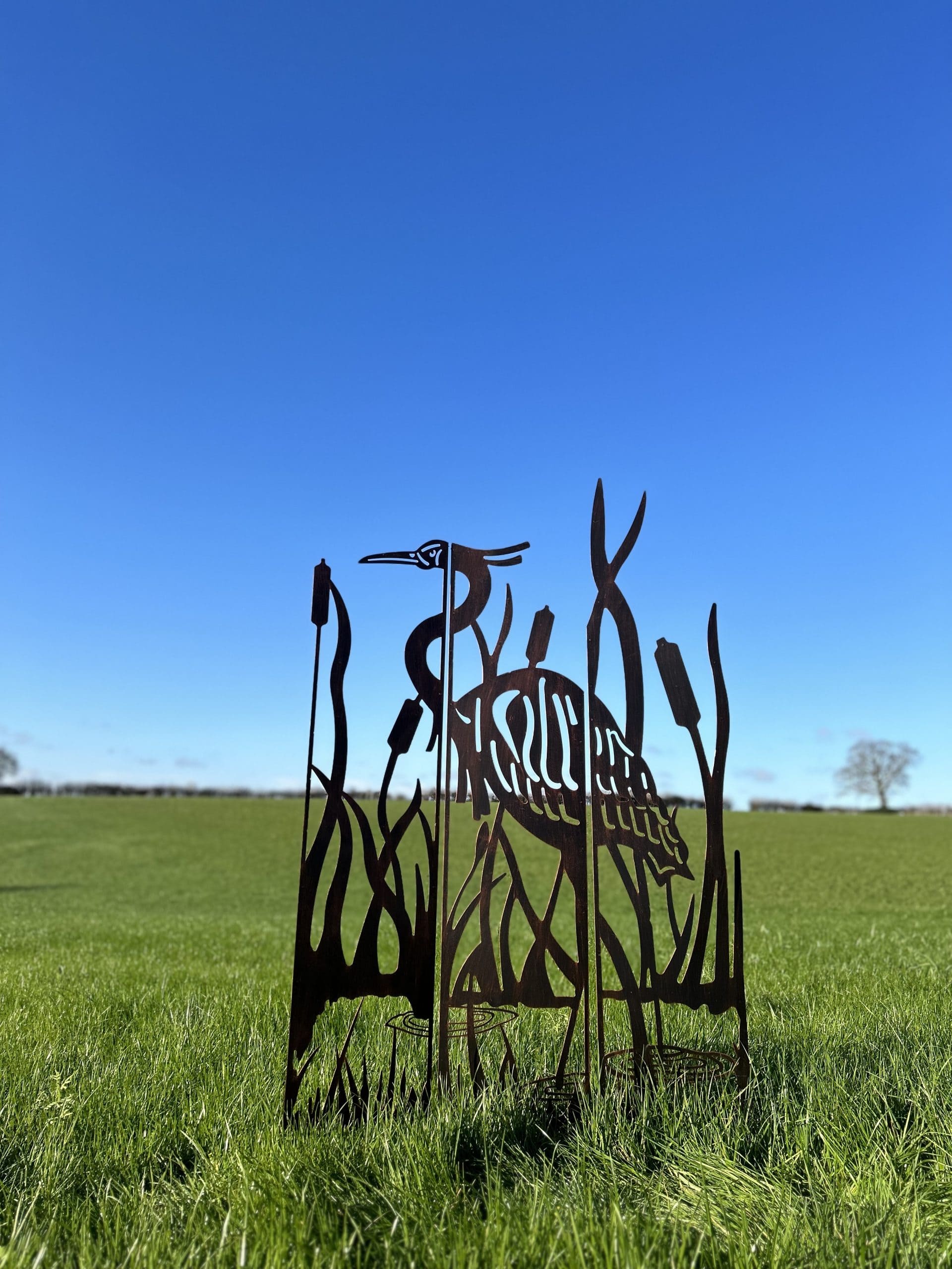 A metal garden sculpture of a heron among reeds, shown outdoors in a grassy field under a clear blue sky.