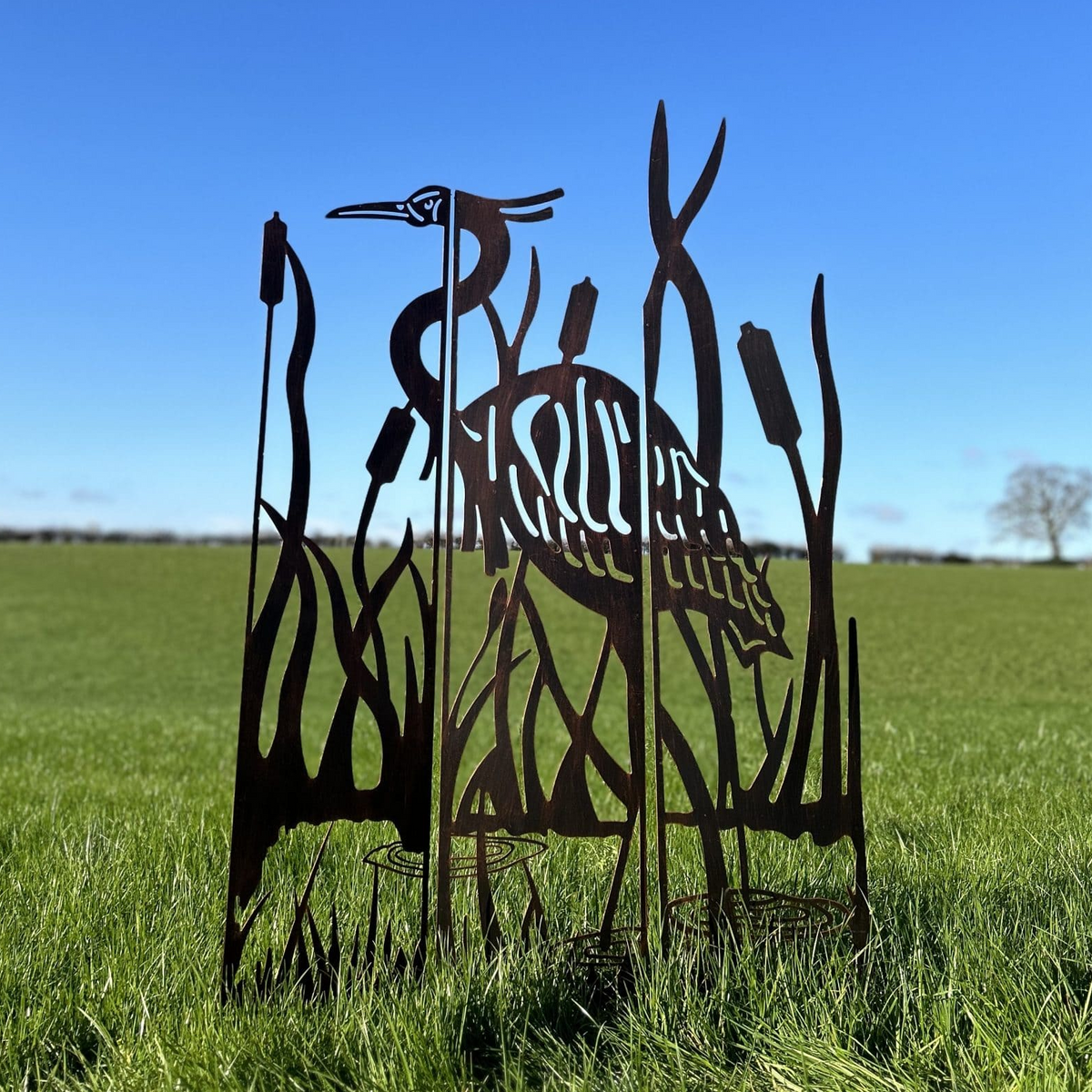 A metal yard sculpture of a heron standing among reeds, photographed outdoors in a grassy field under a clear blue sky.