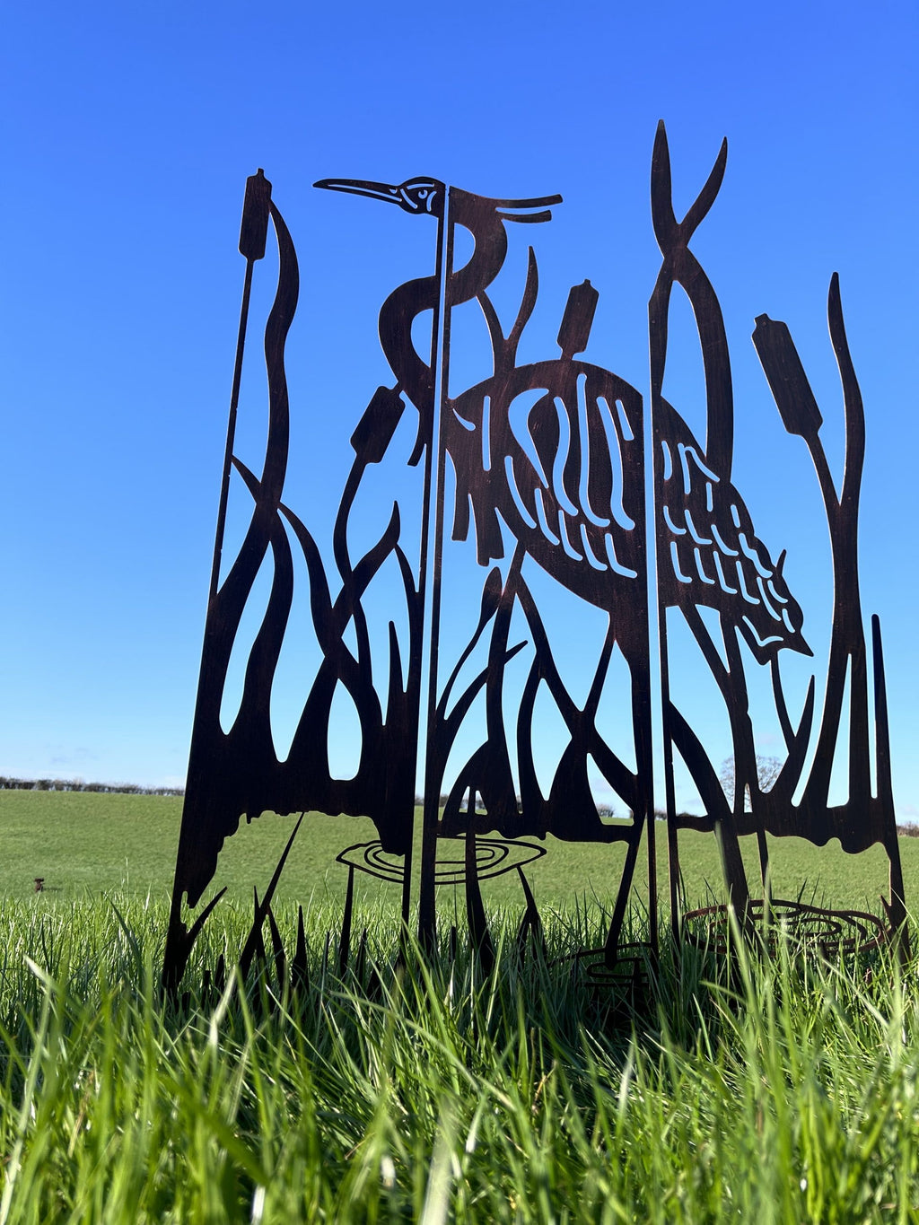 Black metal yard sculpture of a heron among reeds, displayed outdoors in a grassy field under a clear blue sky.