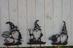 Three black metal garden gnome yard signs with star-patterned hats are displayed against a white vertical wood plank fence.