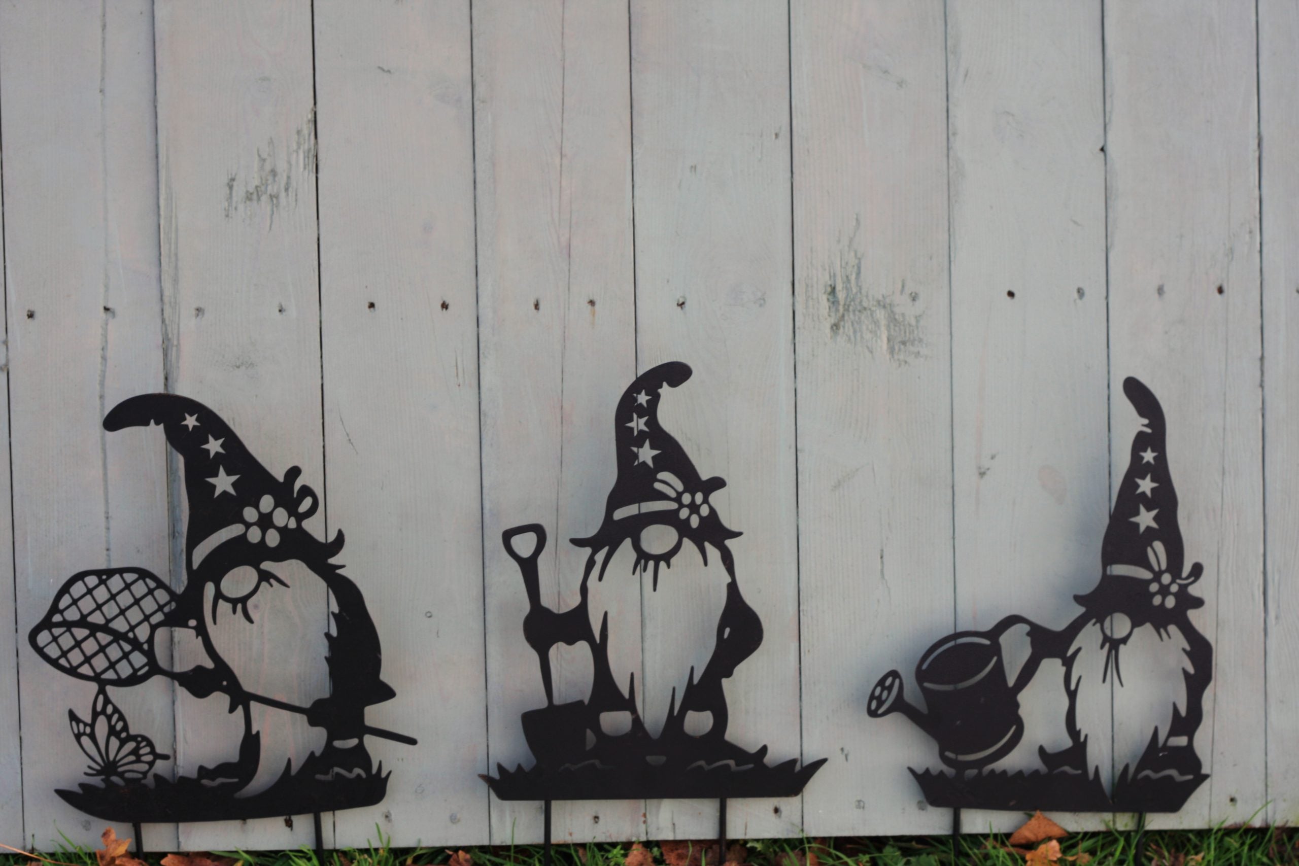 Three black metal garden gnome yard signs with star-patterned hats are displayed against a white vertical wood plank fence.