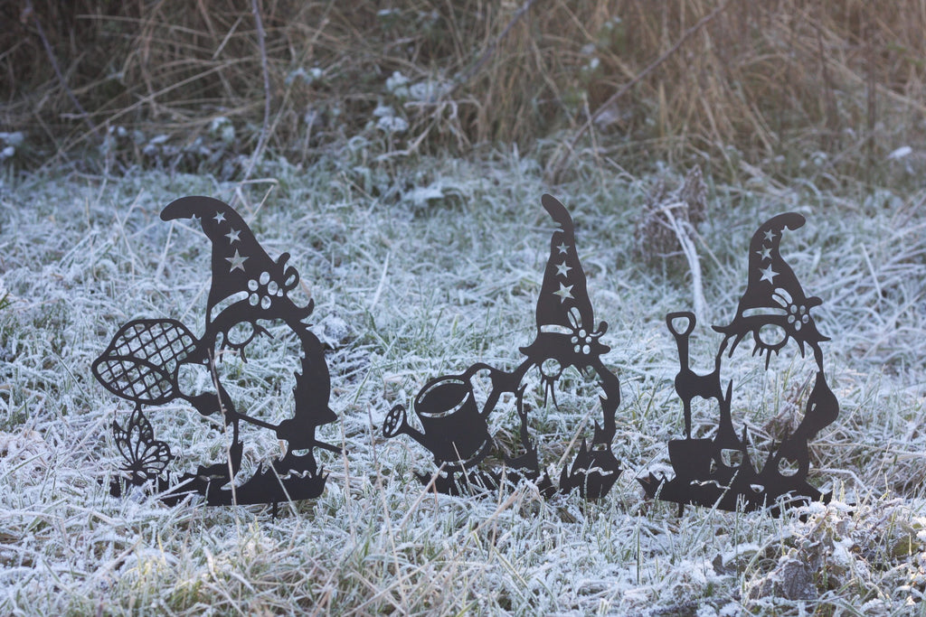 Three black metal garden gnome cutouts with winter-themed accessories, displayed on frosty grass outdoors.