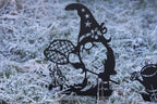 A black metal garden stake shaped like a whimsical witch figure with a pointed hat, net, butterfly, and small cat, displayed outdoors in frosty grass.
