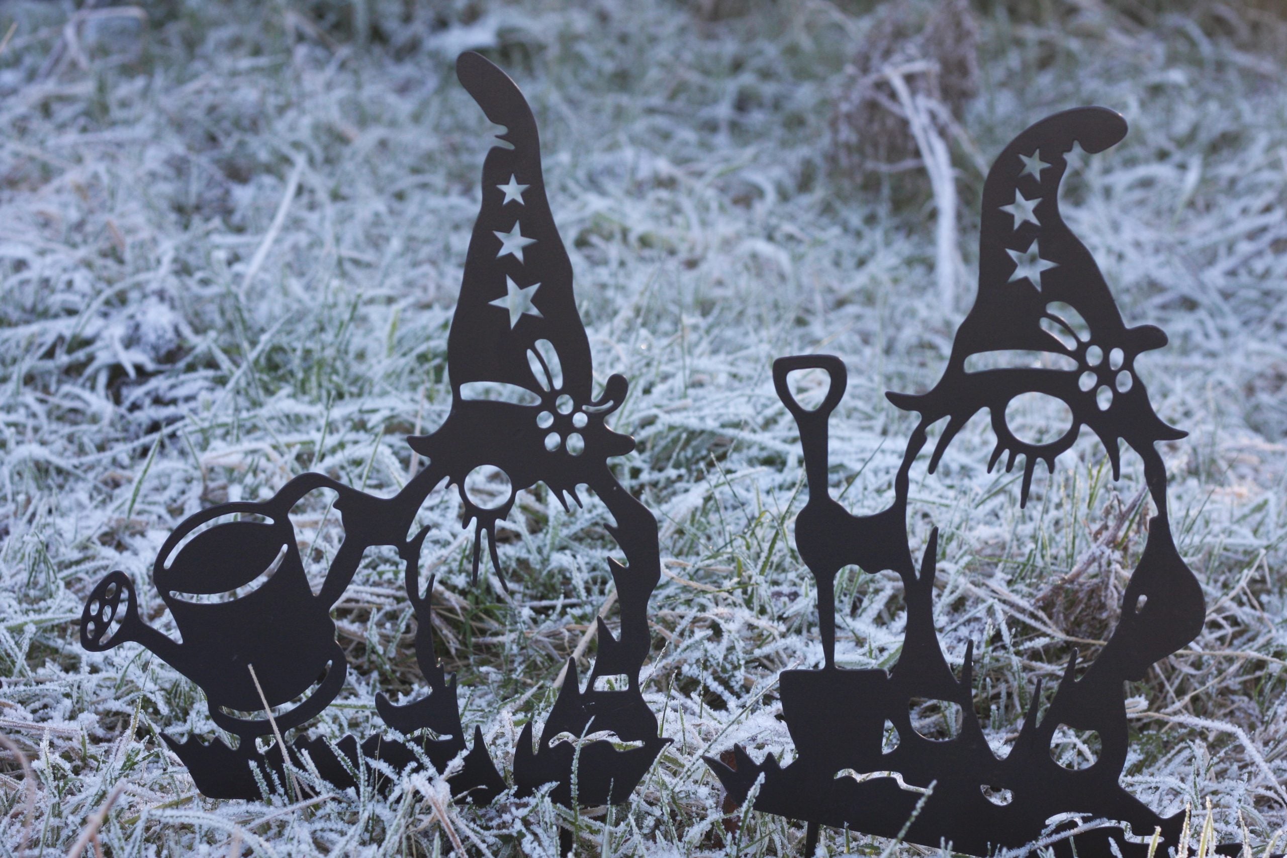 Two black metal garden gnome stakes with star details and accessories, displayed in frosty grass outdoors.