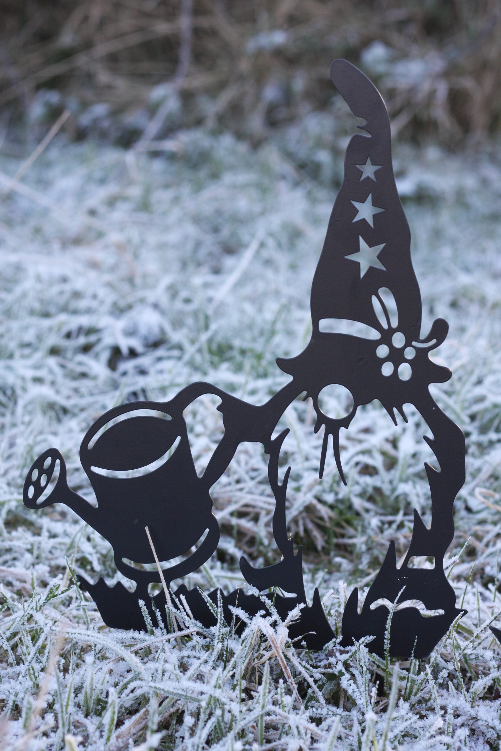 A black metal garden gnome and watering can decoration standing in frosty grass outdoors.
