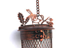A close-up of a hanging metal lantern or cage with a decorative top featuring bird and leaf motifs, shown against a plain white background.