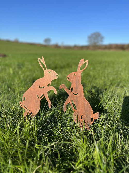 Two small wooden rabbit garden stakes standing in green grass outdoors under a clear blue sky.