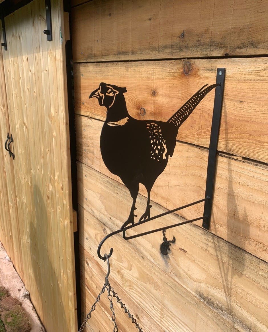 A black metal hanging rack shaped like a pheasant bird, mounted on a wooden wall next to a wooden door, with a hanging chain visible below.