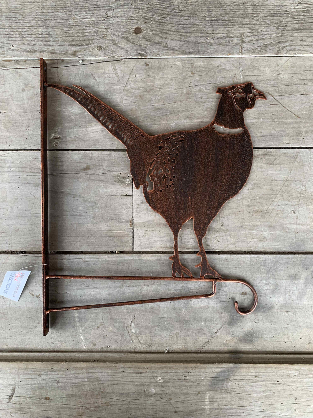Rustic metal wall decor shaped like a pheasant standing on a small hook stand, made from weathered copper-toned metal and mounted on a light gray wood plank background.