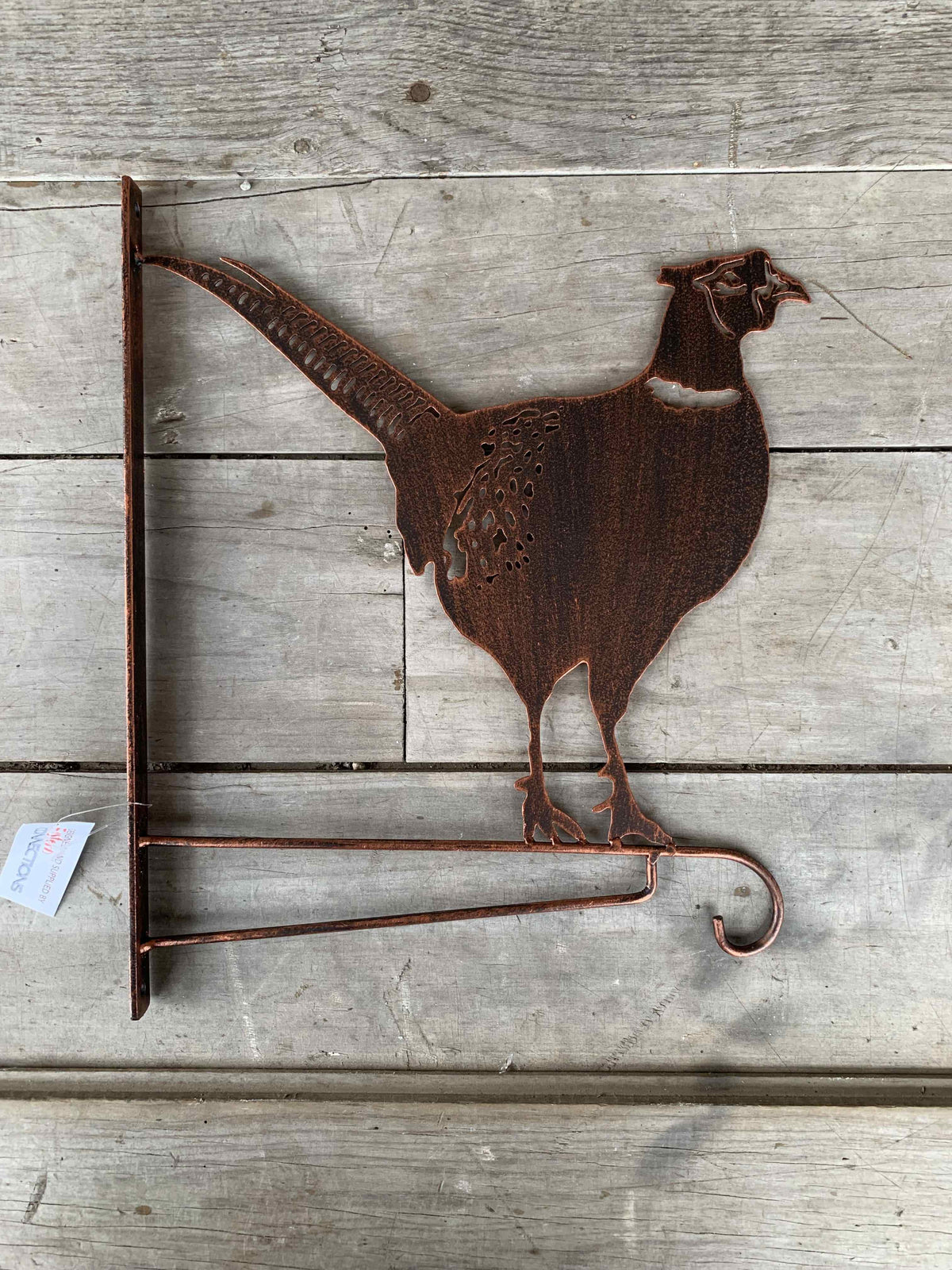 Rustic metal wall decor shaped like a pheasant standing on a small hook stand, made from weathered copper-toned metal and mounted on a light gray wood plank background.