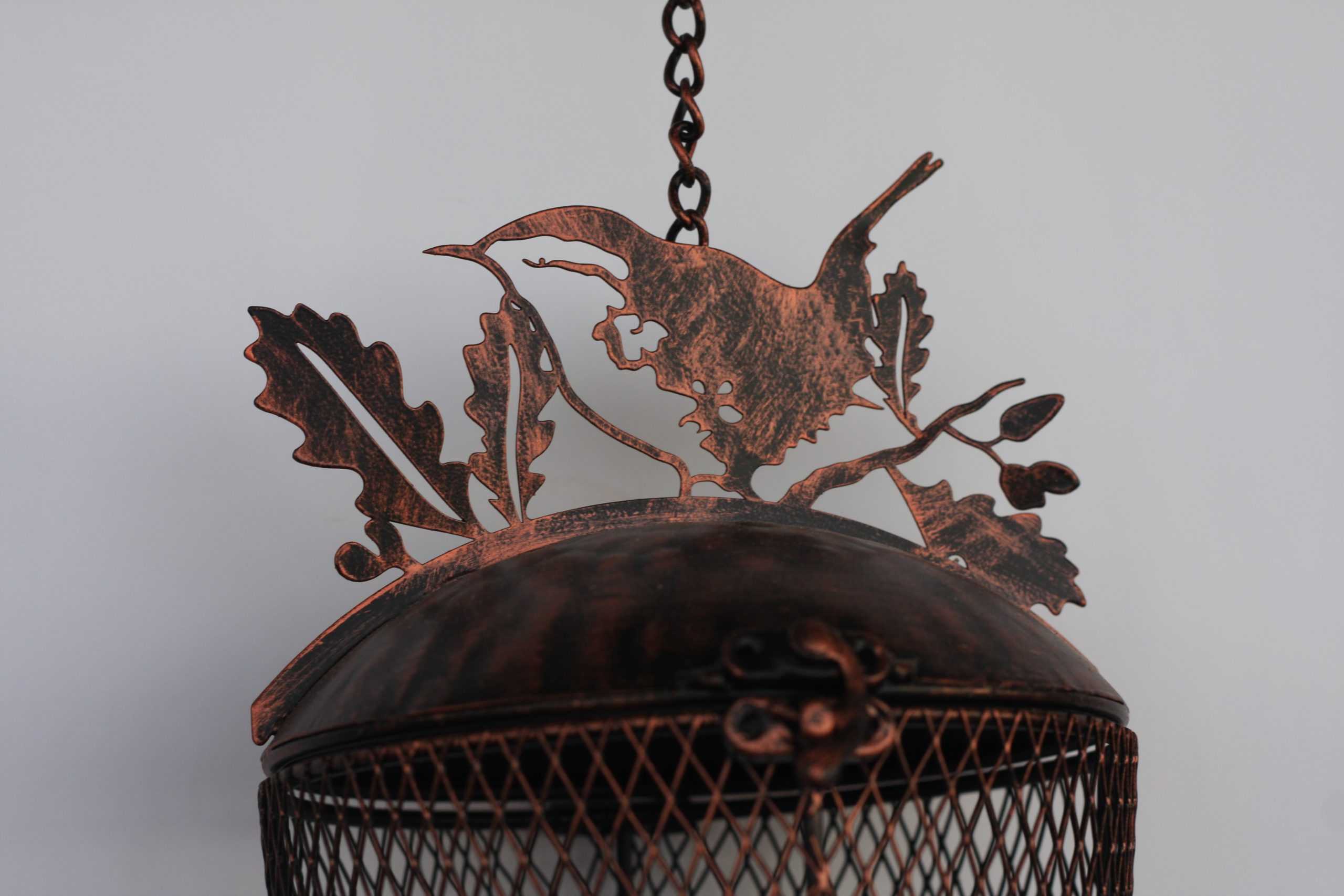 A close-up of a hanging metal lantern or cage with a domed top featuring a cutout bird and oak leaves in a distressed copper finish, suspended by a chain against a plain light gray background. Metal wire basket with a decorative lid held by a hand on a white background bird feeder antique metal steel uk garden decor