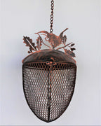 A rustic metal hanging bird feeder with a copper-toned top and a dark mesh-covered shade, suspended by a chain from a hook against a plain light background.