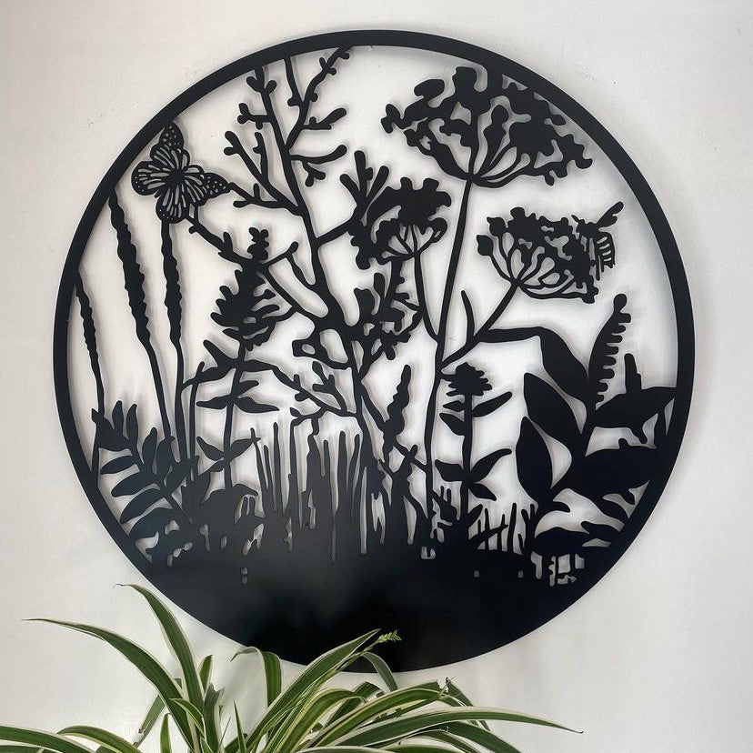 Black metal circular wall art featuring a botanical silhouette design with a butterfly and wildflowers, mounted on a white wall above a green plant.