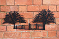 Black metal wall art featuring two stylized trees and a fence, mounted on a red-orange brick wall.