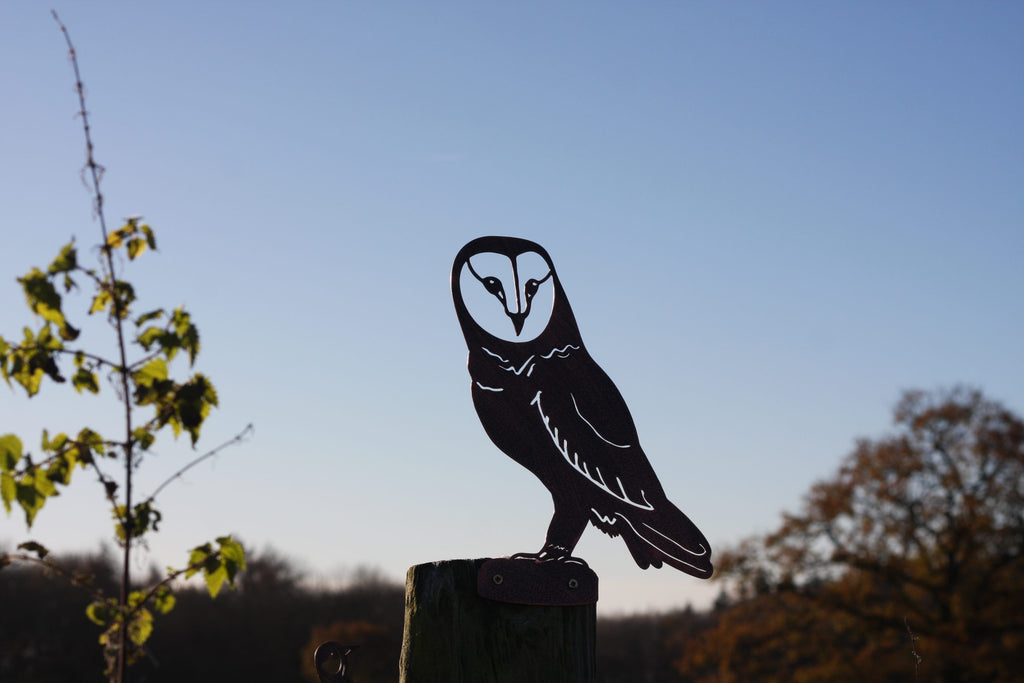 A black metal owl silhouette cutout mounted on a wooden post outdoors, with a blue sky and blurred trees in the background.