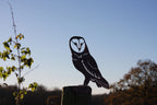 A black metal owl silhouette cutout mounted on a wooden post outdoors, with a blue sky and blurred trees in the background.