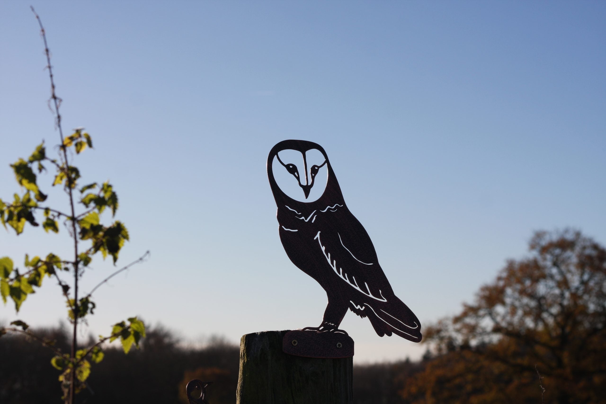 A black metal owl silhouette cutout mounted on a wooden post outdoors, with a blue sky and blurred trees in the background.
