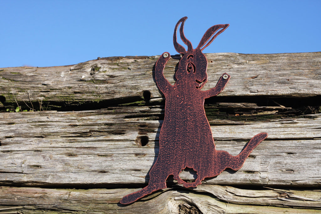 A rusted metal Easter bunny silhouette wall decor piece mounted on a weathered wooden fence under a clear blue sky.