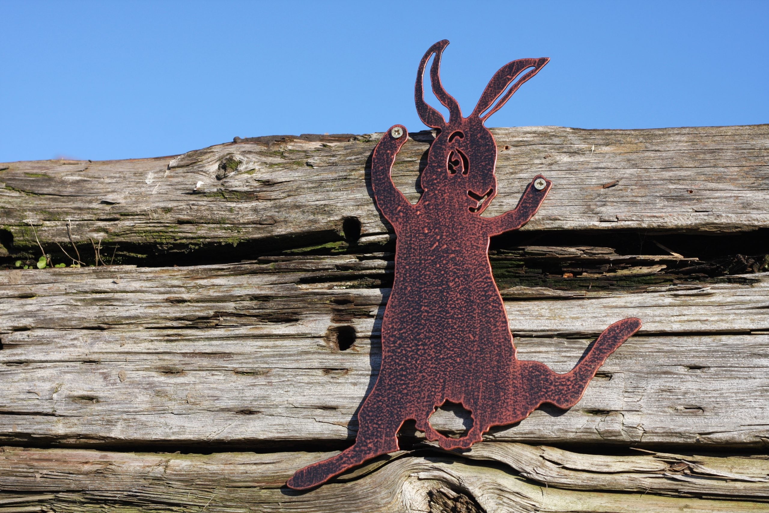 A rusted metal Easter bunny silhouette wall decor piece mounted on a weathered wooden fence under a clear blue sky.