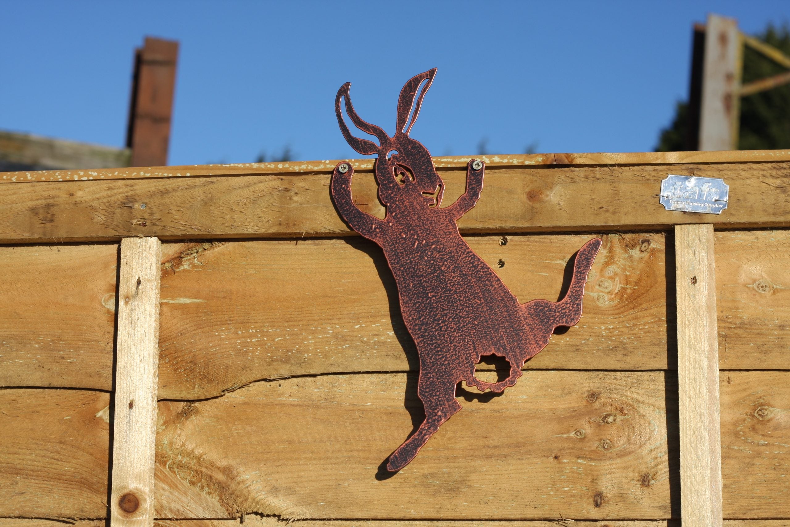 A rusted metal hare silhouette wall decor piece mounted on a wooden fence against a blue sky.