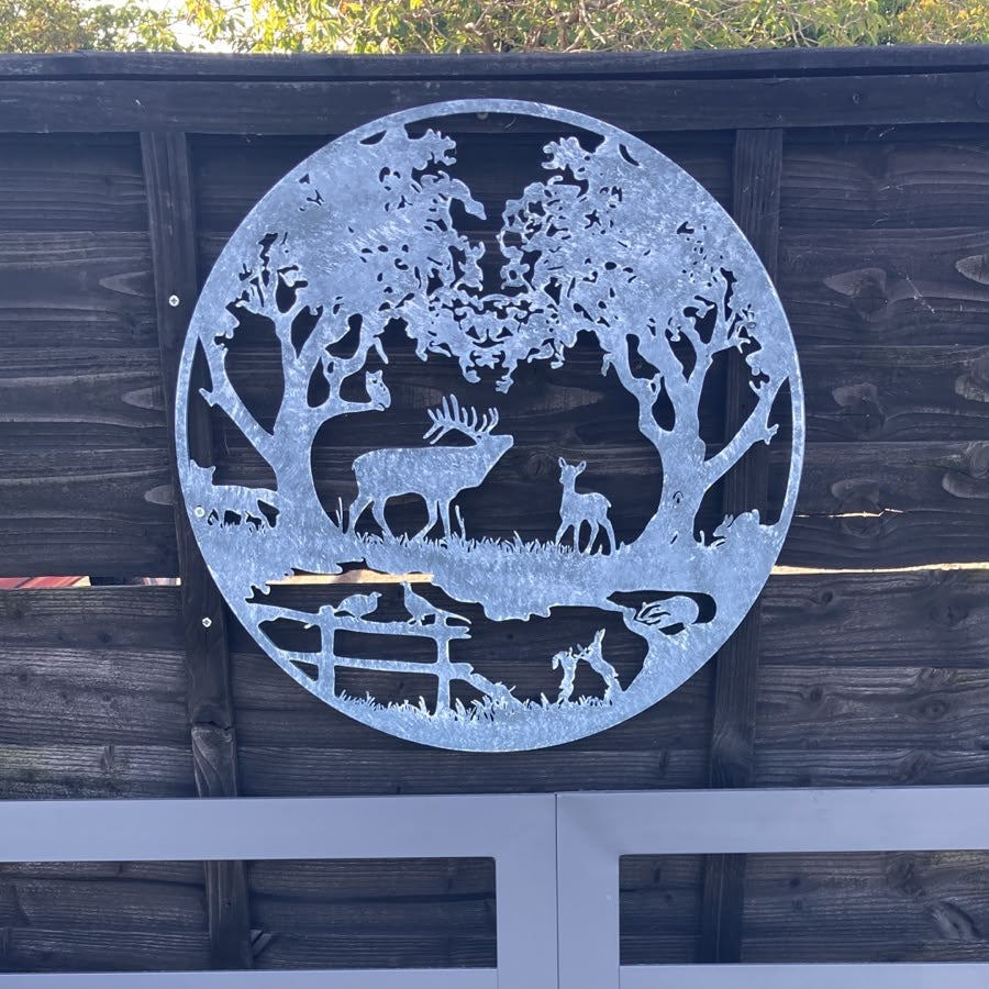 A round metal wall art piece featuring a forest scene with deer and a bench, mounted on a dark wooden fence above a bed with blue bedding.