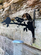 Black metal bird feeder wall mount featuring silhouettes of three birds on a branch, shown mounted on a rustic weathered wood wall with a hanging wire bird feeder in front.