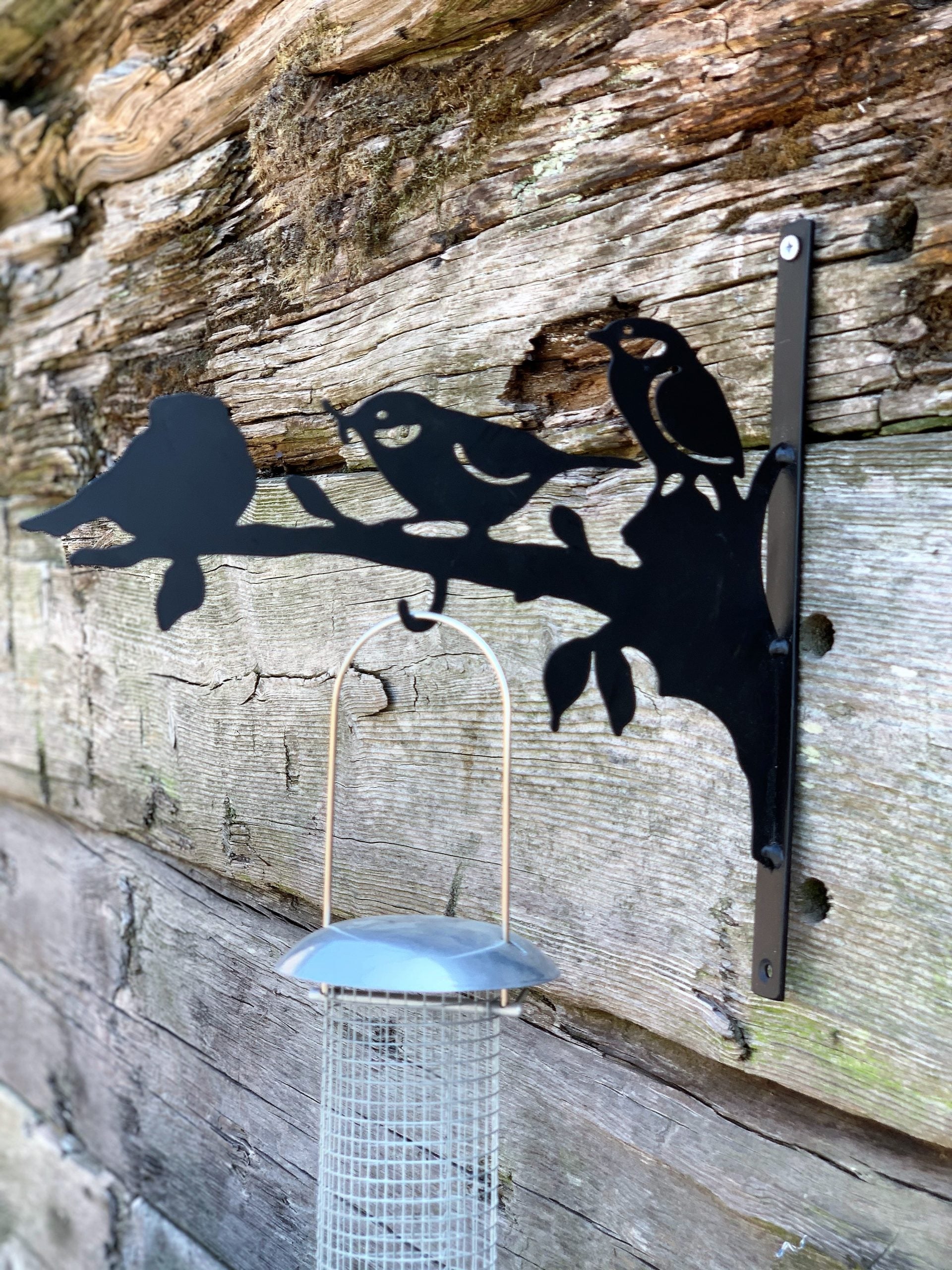 Black metal bird feeder wall mount featuring silhouettes of three birds on a branch, shown mounted on a rustic weathered wood wall with a hanging wire bird feeder in front.