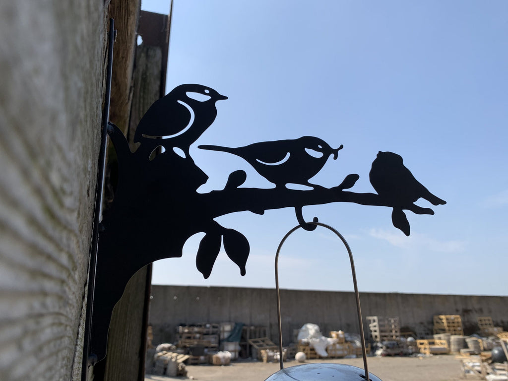 Black metal bird silhouette wall decor mounted on a wooden post outdoors, with three birds perched on a branch and a hanging loop above.