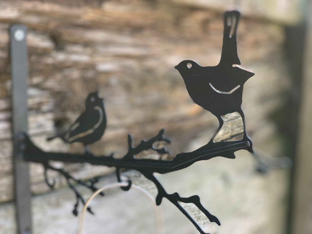 Black metal bird perched on a branch wall hanging with two birds and branch silhouettes, shown against a blurred rustic wood background.