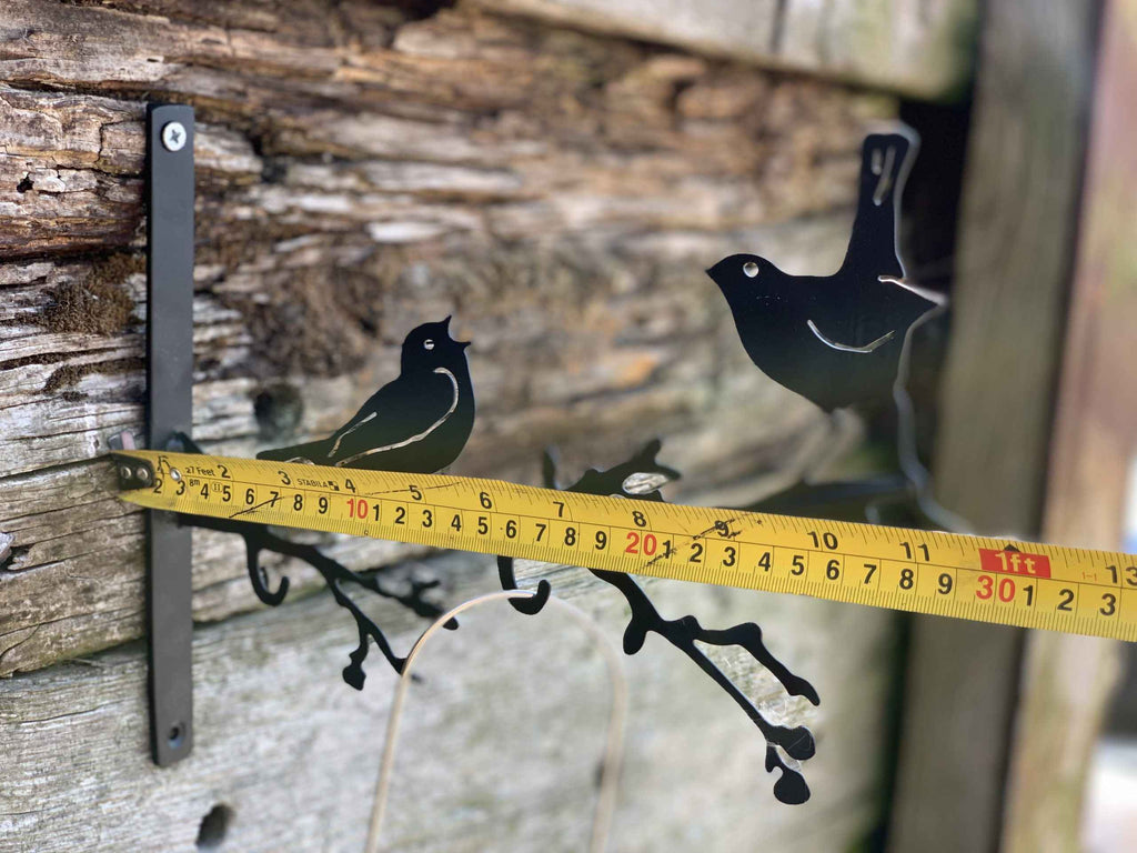 Black metal bird perched on a branch garden stake with a yellow tape measure showing the height against a rustic wooden wall.