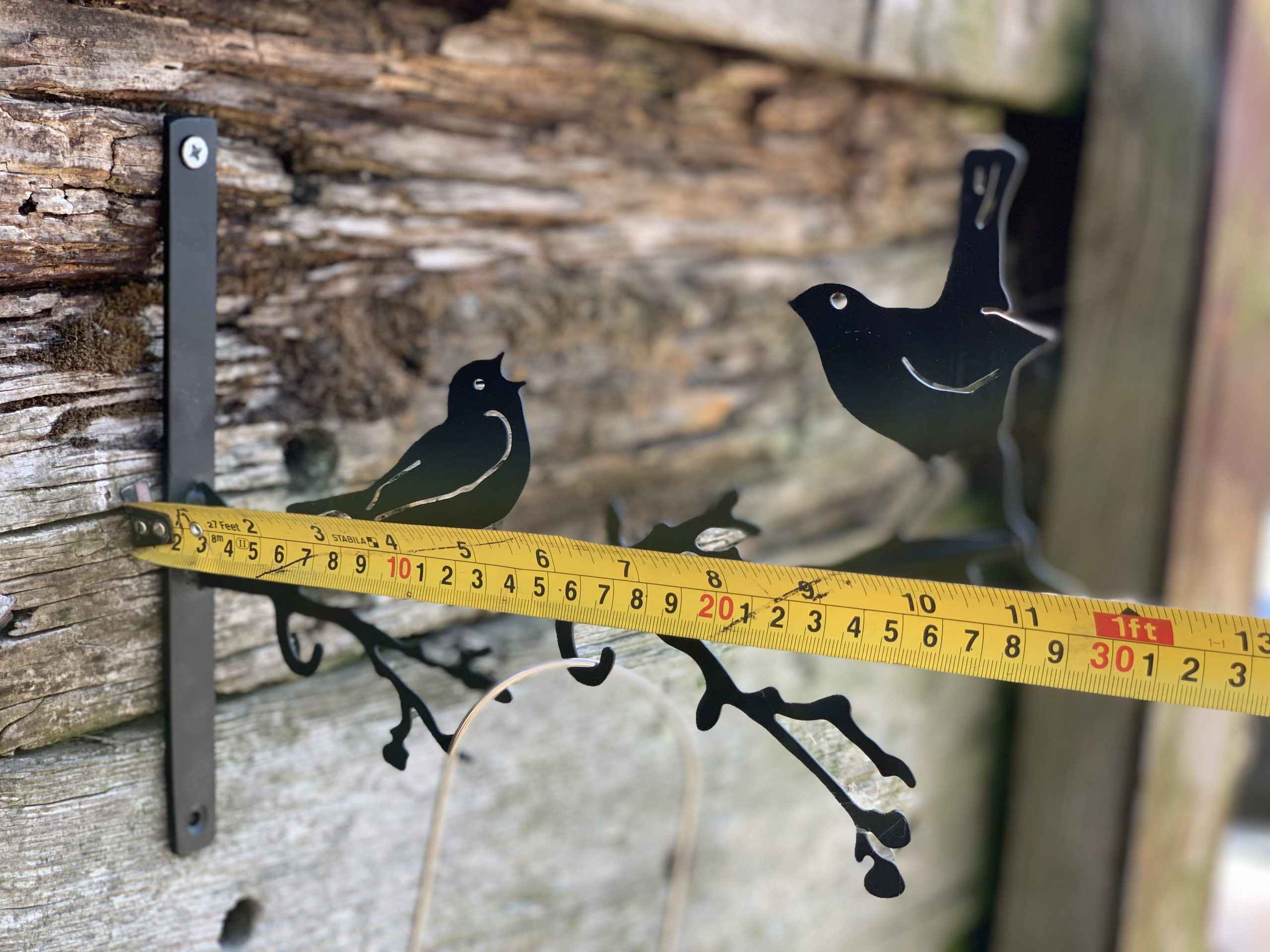 Black metal bird perched on a branch garden stake with a yellow tape measure showing the height against a rustic wooden wall.