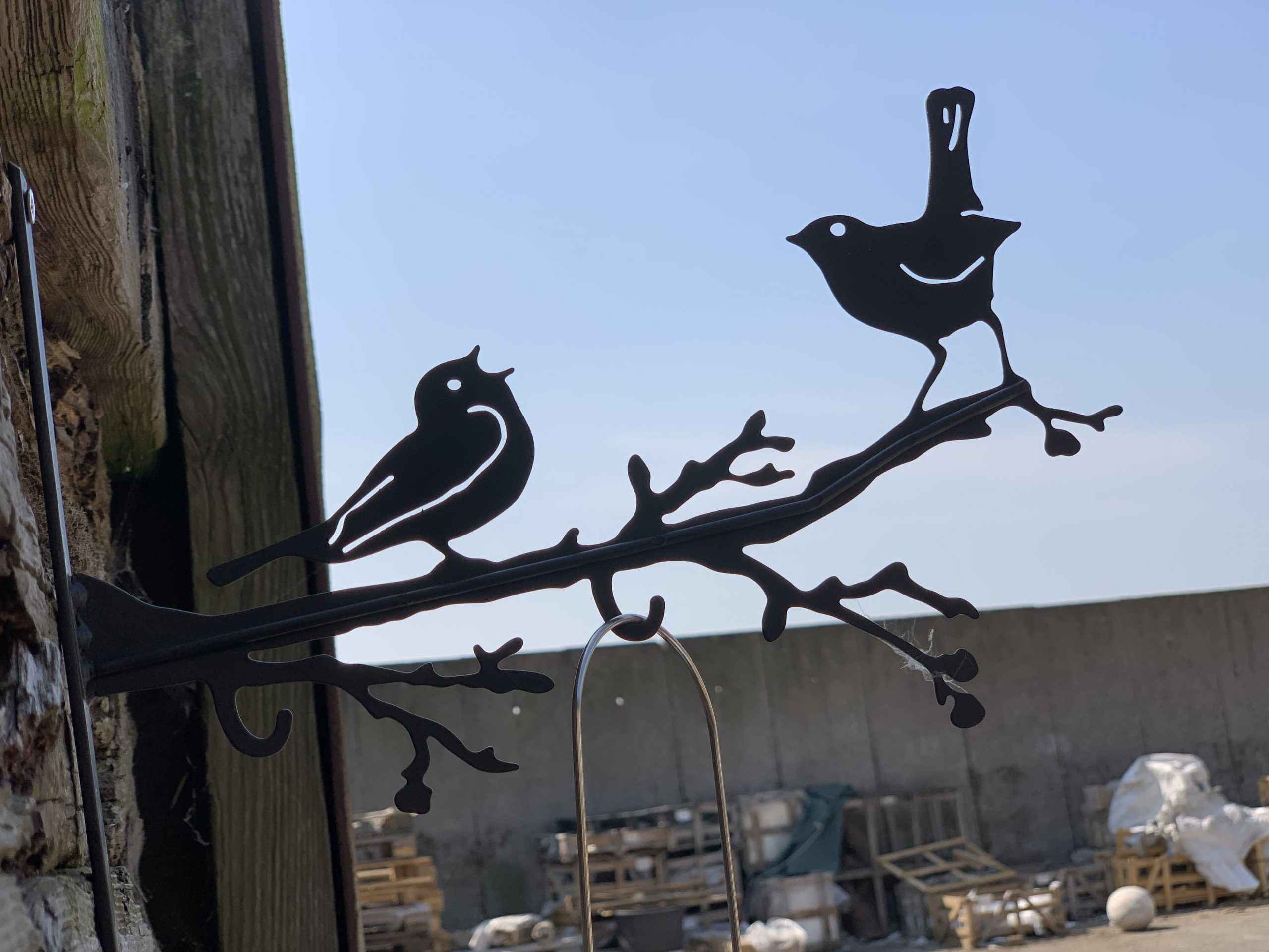 Black metal bird perched on branch wall hanging with two birds and a curved hook, shown mounted on a wooden post outdoors with a concrete wall and sky behind it.