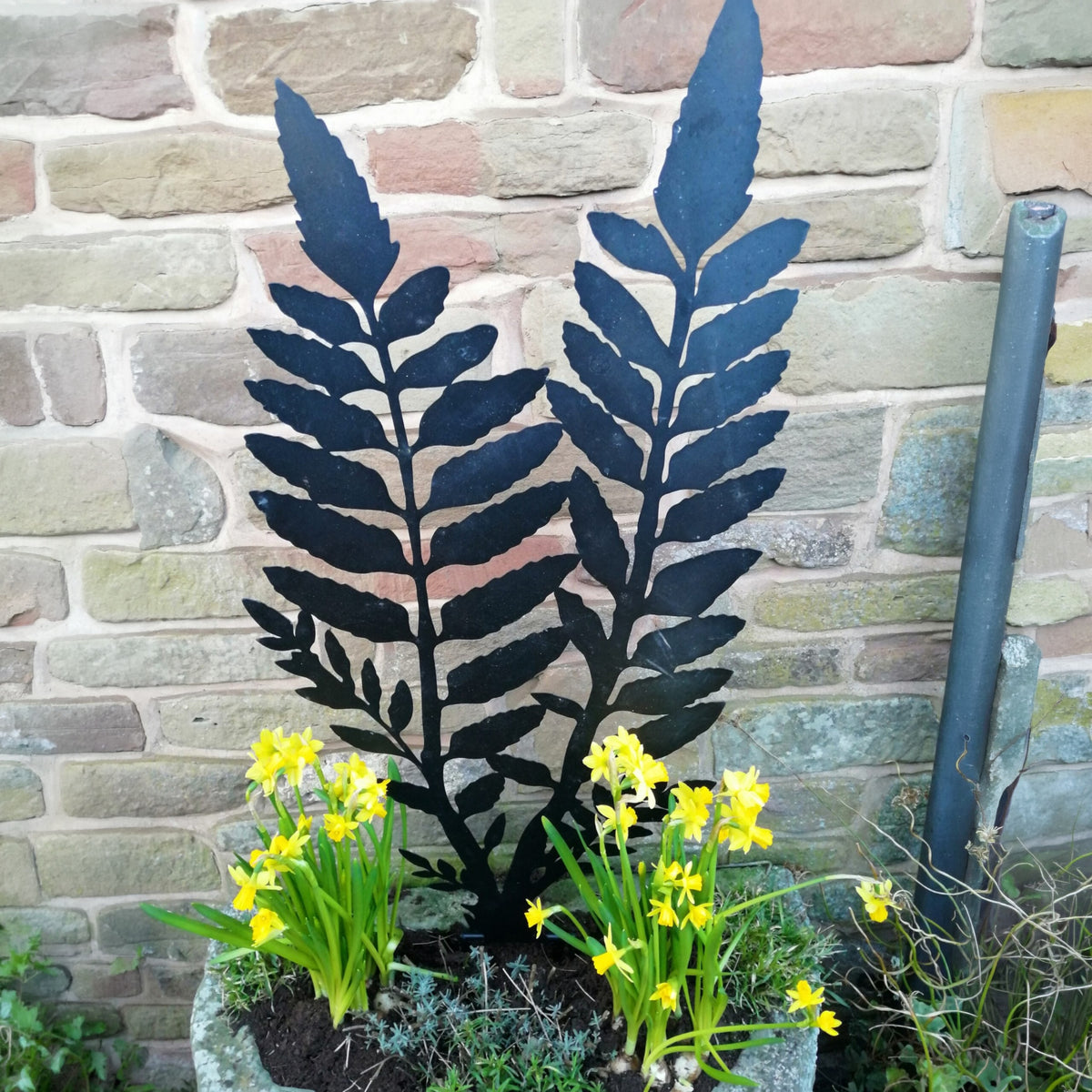 Black metal fern wall art displayed outdoors in a garden planter with yellow daffodils in front of a stone brick wall.