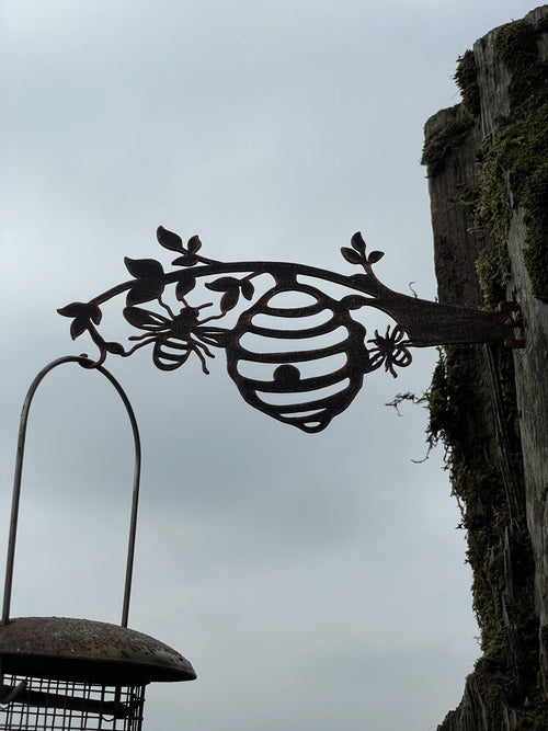 A dark metal outdoor sign shaped like a beehive on a branch with leaf and bee motifs, mounted on a weathered wooden post against an overcast sky. A bird feeder is partially visible at the lower left.