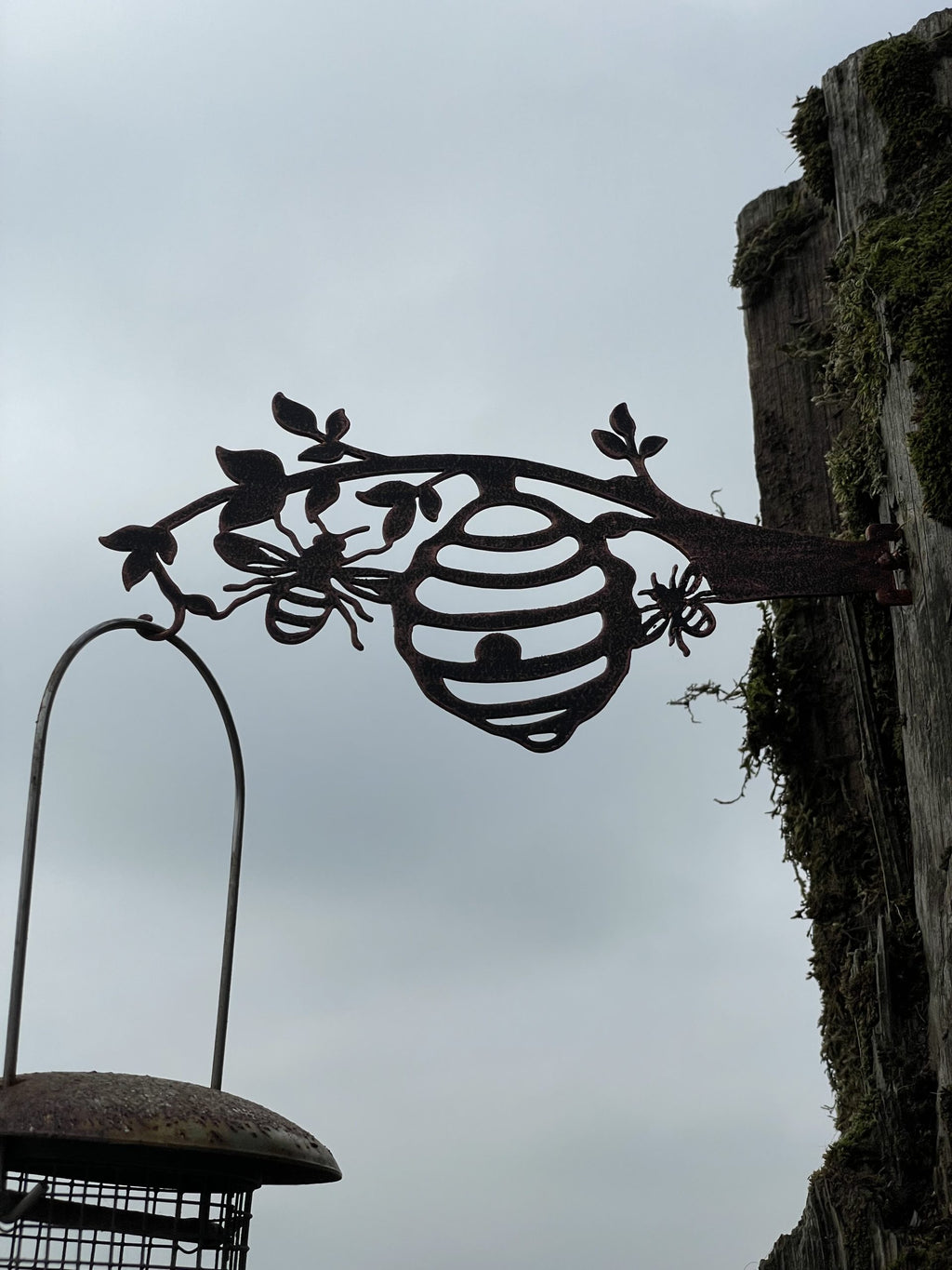 A dark metal outdoor sign shaped like a beehive on a branch with leaf and bee motifs, mounted on a weathered wooden post against an overcast sky. A bird feeder is partially visible at the lower left.
