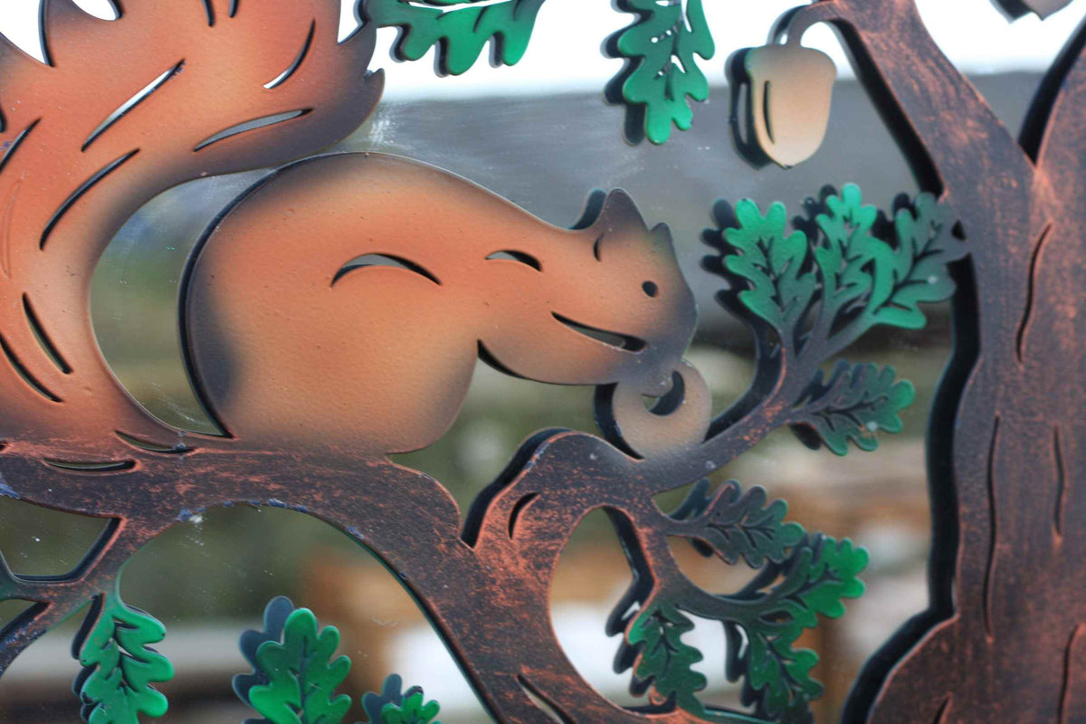 A close-up of a decorative metal cutout featuring a smiling squirrel nestled among brown tree branches and green leaves, with a small hanging acorn at the top right.