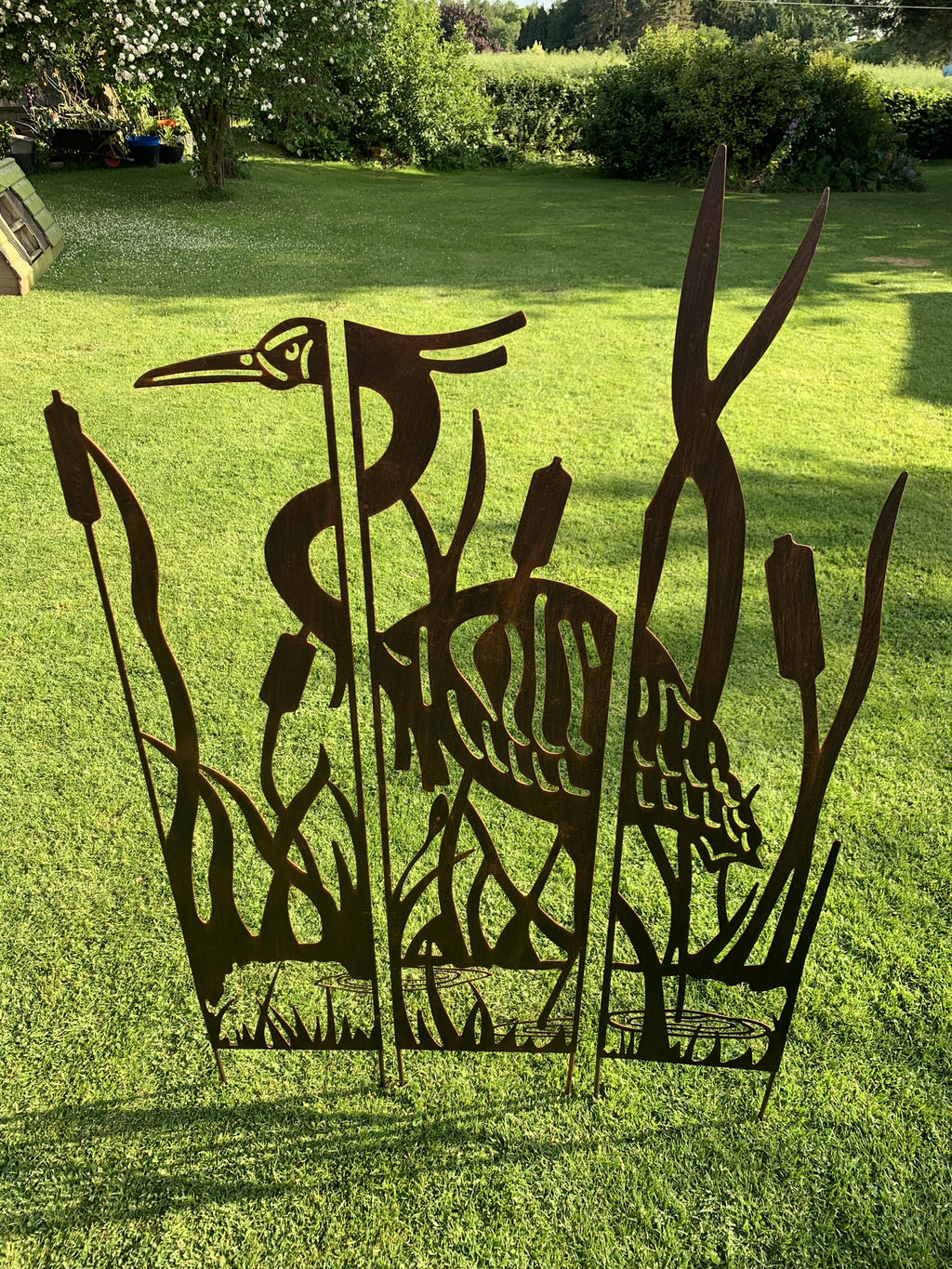 A dark metal outdoor garden sculpture featuring a heron among reeds, displayed on a green grass lawn in a backyard setting.