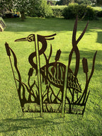A dark metal outdoor garden sculpture featuring a heron among reeds, displayed on a green grass lawn in a backyard setting.