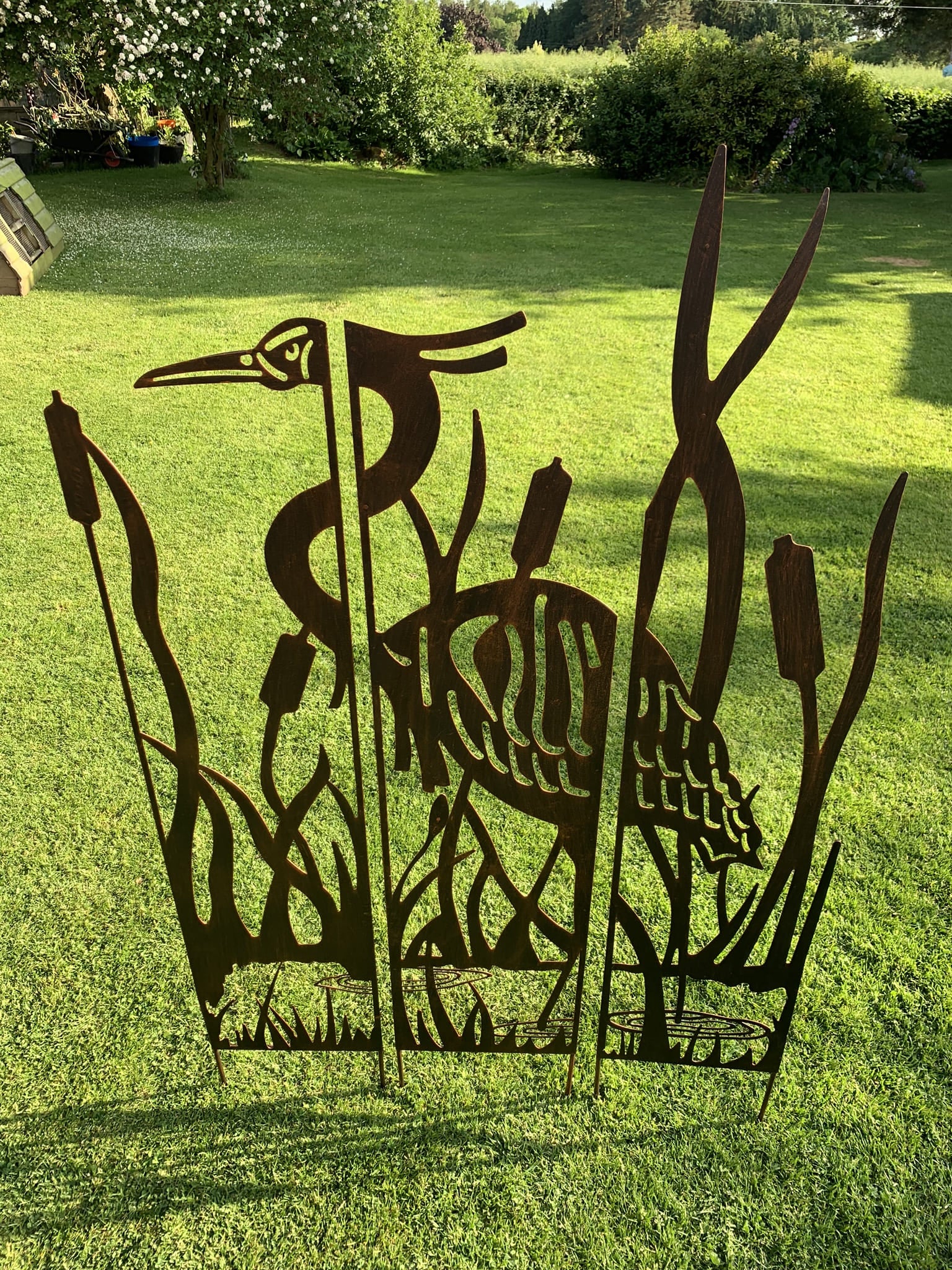 A dark metal outdoor garden sculpture featuring a heron among reeds, displayed on a green grass lawn in a backyard setting.
