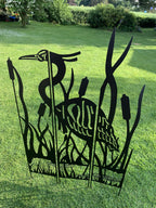 Black metal yard sculpture of a heron standing among reeds, displayed on a green grass lawn outdoors.