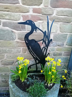 A black metal garden sculpture of a heron with reeds, displayed in a stone planter with yellow daffodils in front of a rustic stone wall.