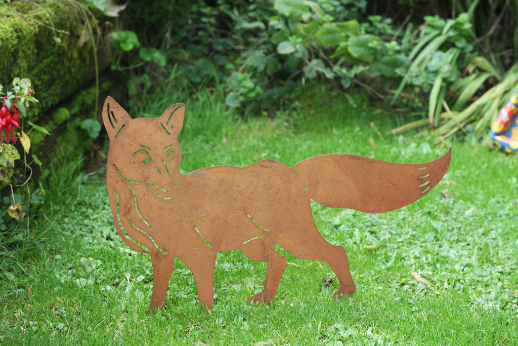 A rust-colored metal cutout of a fox standing on a green grassy lawn with garden foliage in the background.