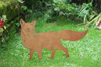 A rust-colored metal cutout of a fox standing on a green grassy lawn with garden foliage in the background.