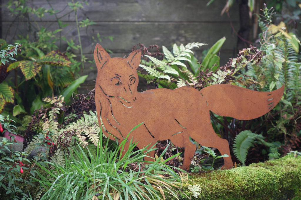 A rusted metal fox silhouette garden stake stands in a lush green garden bed with ferns and moss.